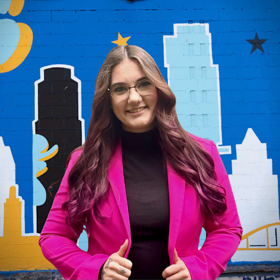 A woman with long hair and glasses is wearing a pink blazer and a black top, standing in front of a colorful mural.