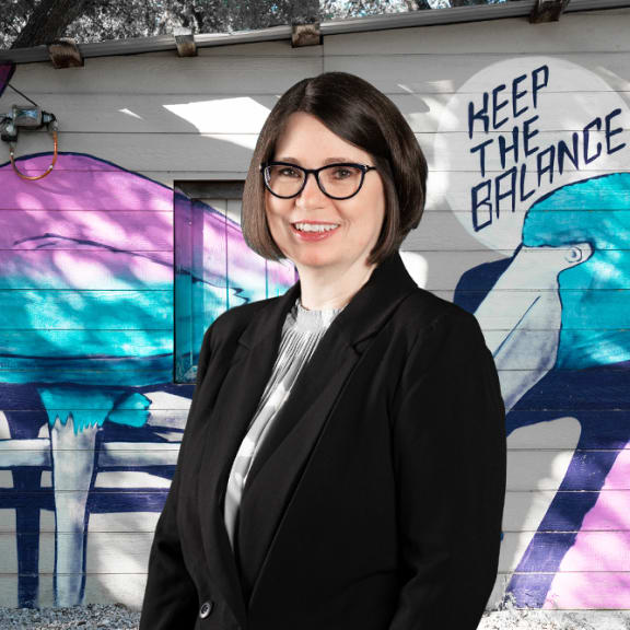 a woman in a suit and glasses stands in front of a mural that says keep the balance