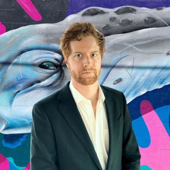 A man in a suit stands in front of a painting of a shark.