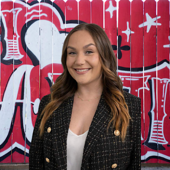 A woman with a white top and black and white checkered jacket standing in front of a red wall with white graffiti.