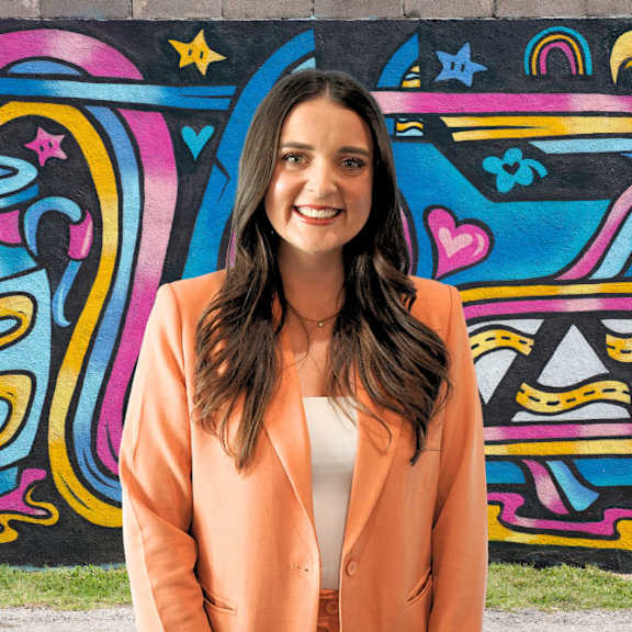 A woman in an orange blazer stands in front of a colorful wall.