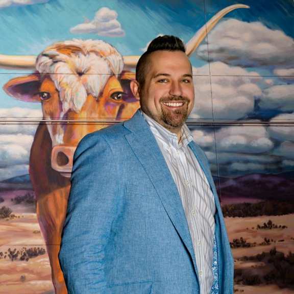 A man in a blue suit stands in front of a painting of a cow.