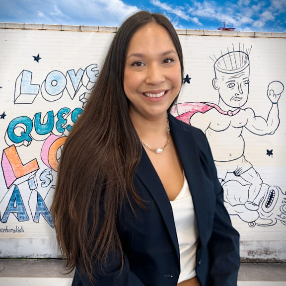 A woman with long hair and a black blazer stands in front of a wall with a drawing of a muscular man.