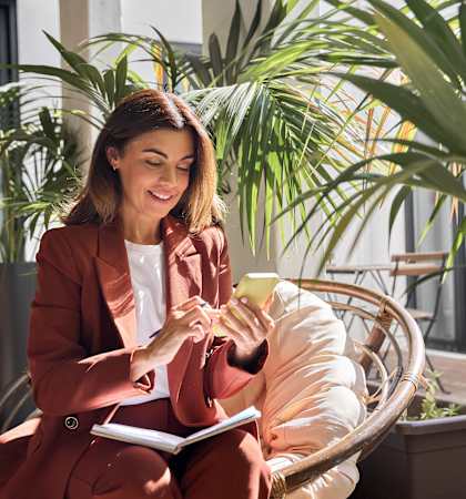A woman in a red suit is sitting on a chair outside, holding a cup of coffee and looking at her phone.