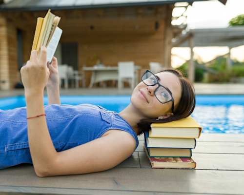 A woman is reading a book while lying on a poolside table.