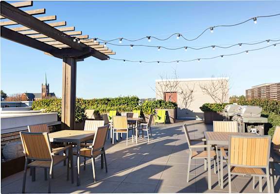 A patio with chairs and tables is covered with a roof and has lights strung above it.