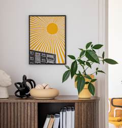 A sunny yellow and black picture hangs on a white wall above a shelf with books, sculptures, and a potted plant.