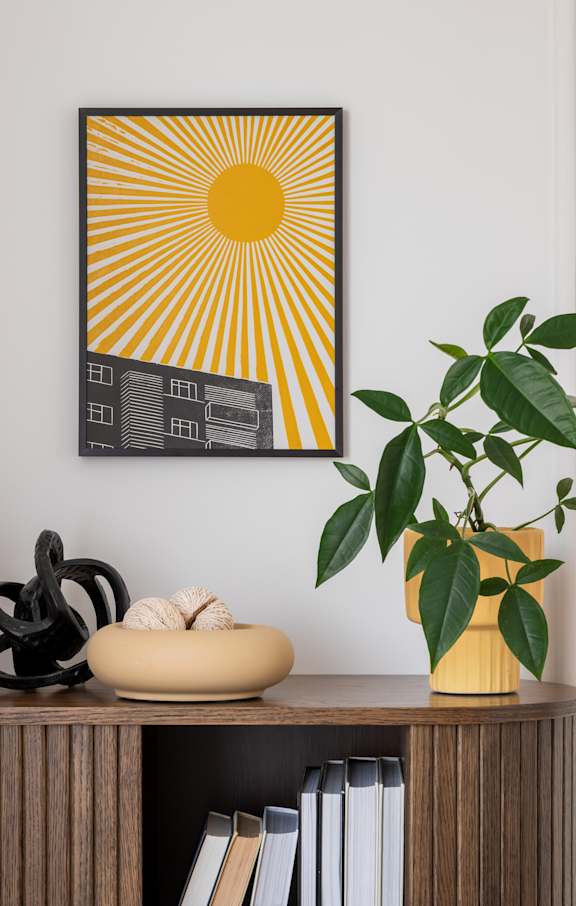 A sunny yellow and black picture hangs on a white wall above a shelf with books, sculptures, and a potted plant.