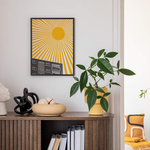 A sunny yellow and black picture hangs on a white wall above a shelf with books, sculptures, and a potted plant.