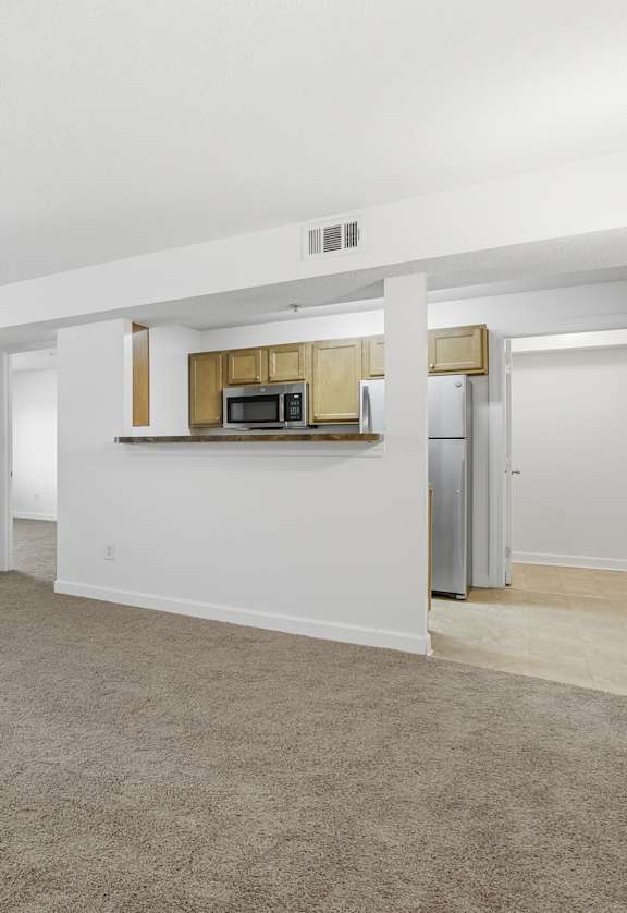 A spacious living room with a carpeted floor and a kitchenette in the background.