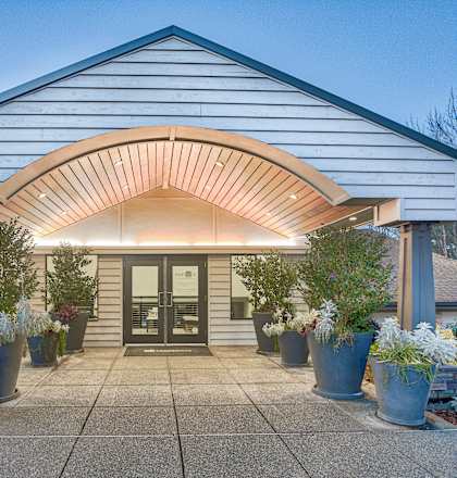 A white building with a black door and a wooden overhang is surrounded by potted plants.