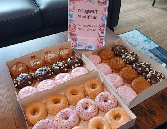 A box of assorted donuts is displayed in front of a sign that says "DOUGHNUT MIND if I do Treat yourself it's Friday after all".