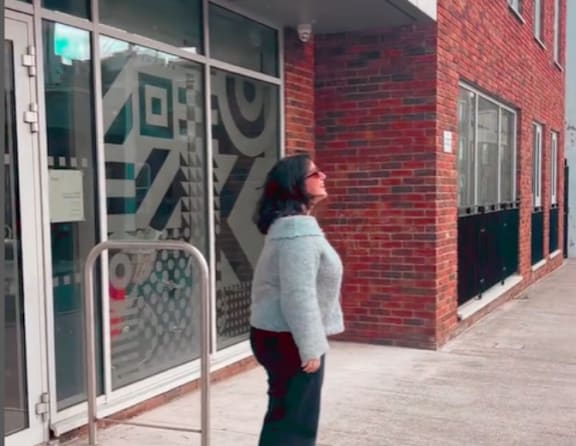 A woman in a grey sweater and red skirt stands in front of a glass door with a red brick building behind her.