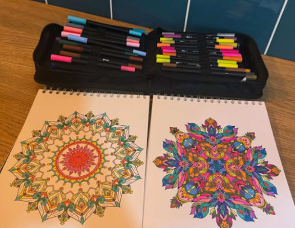 A set of markers is placed on a table next to two pages of colorful mandala drawings.