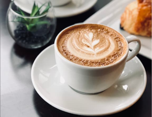 A cup of coffee with a leaf design on top sits on a saucer.