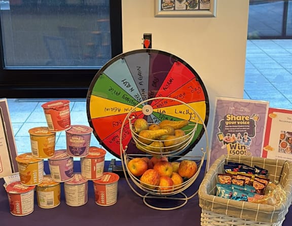 A wheel of fortune is on a table with snacks and a sign that says "Share your voice and win £500!".