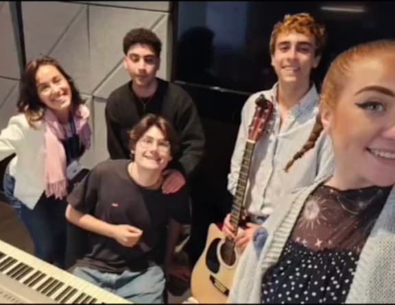 A group of five people are standing together, with one person holding a guitar.