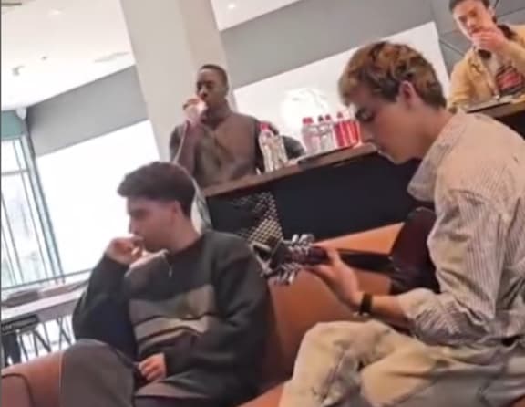 A man is sitting on a couch playing a guitar while another man stands beside him.