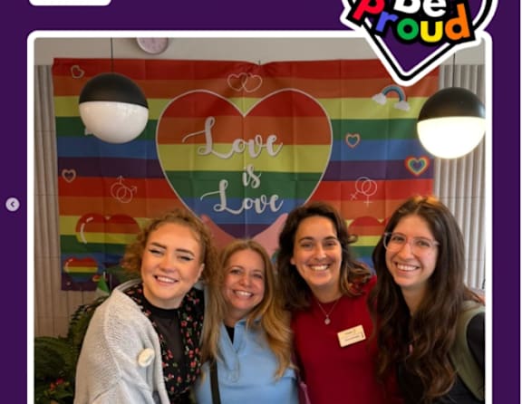 Four people are standing in front of a colorful backdrop with the words "Love is Love" written on it.