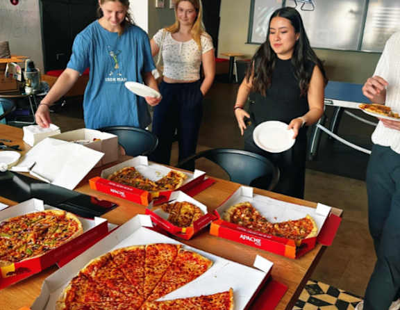 Three women are standing around a table with pizza boxes on it.