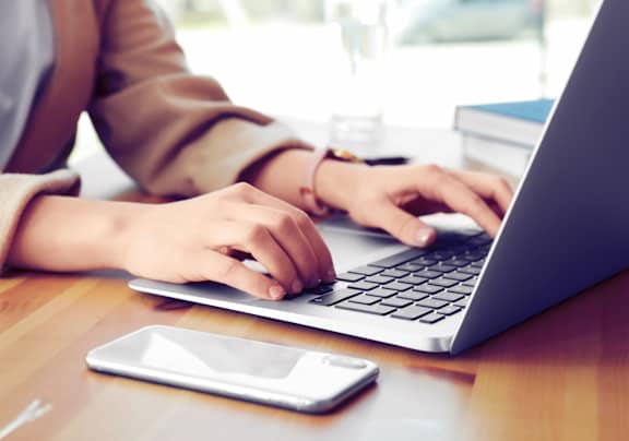 A person is typing on a laptop with a smartphone and glasses on the table.