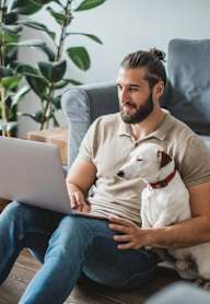 A man is sitting on the floor with a laptop, surrounded by cardboard boxes and a dog is resting on his lap.