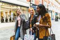 Three women are walking down the street with shopping bags and coffee.