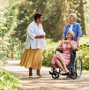 A woman in a yellow skirt is standing on a path in the woods with two other women, one of whom is in a wheelchair.