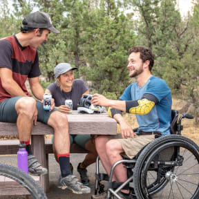 two men sitting at a picnic table with a man in a wheelchair
