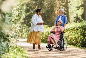 A woman in a yellow skirt is standing on a path in the woods with two other women, one of whom is in a wheelchair.