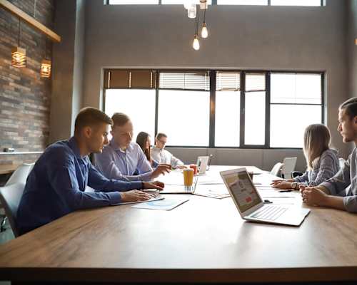 A group of people are sitting around a table with laptops and papers, appearing to be in a meeting.