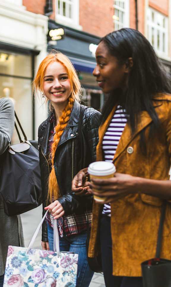Three women are walking down the street with shopping bags and coffee.