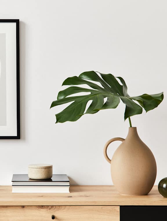 A framed artwork hangs on a wall above a desk with a chair, a plant, and some books.