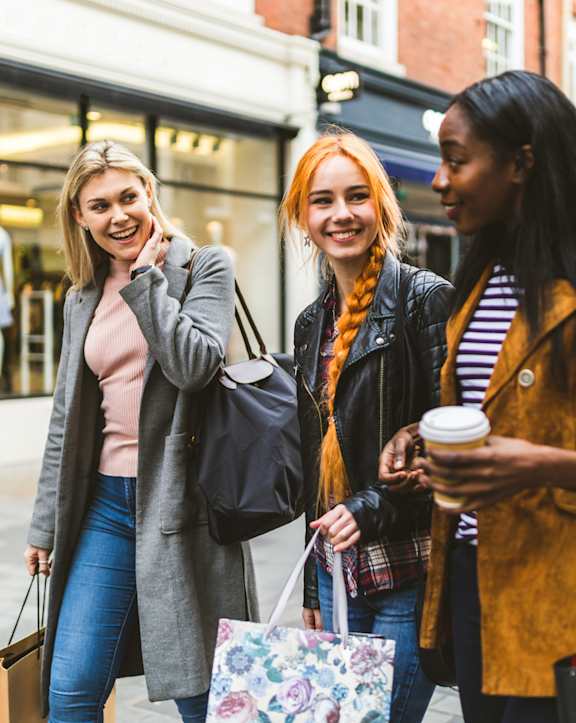 Three women are walking down the street with shopping bags and coffee.