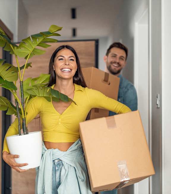 A woman is holding a potted plant and a cardboard box while a man is carrying another cardboard box.