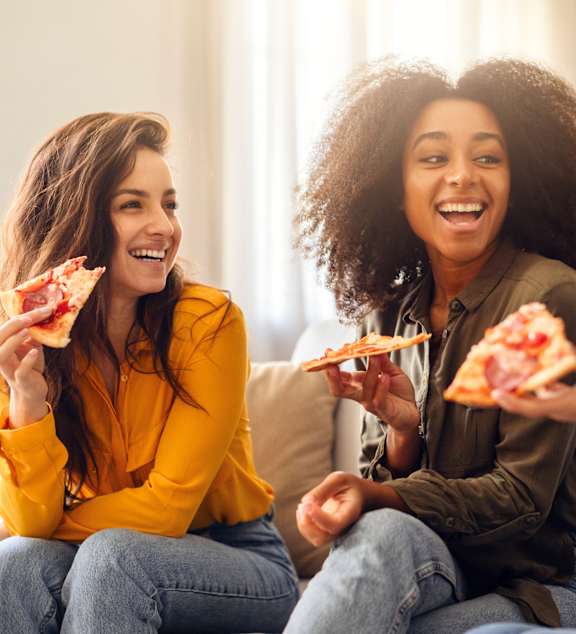 Three women are sitting on a couch eating pizza.