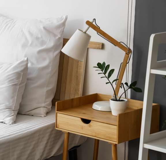 A bed with white pillows and a wooden nightstand with a lamp and plant.