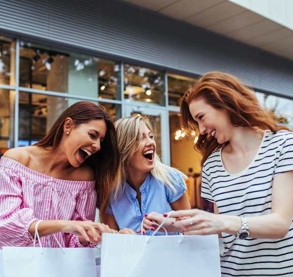 Three women are laughing and looking at a phone while holding shopping bags.at The Liliana Apartment Homes, Pooler Georgia