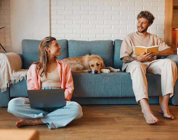 A man and woman sitting on the floor in front of a couch with a dog between them, the woman is using a laptop and the man is reading a book.