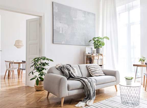 Kerrville, TX, Apartments Near Peterson Regional Medical Center - Winwood Club - Sunlit Living Room With Gray Sofa, Indoor Plants, Wood Floors, and Large Windows With Sheer Curtains.