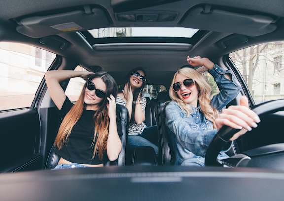 Three women wearing sunglasses are sitting in a car and laughing.