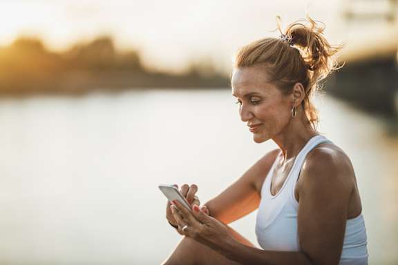 A woman in a white tank top is sitting by the water, looking at her phone.