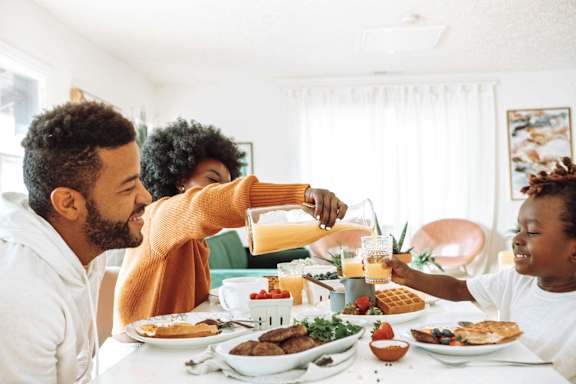 A man pours juice into a glass while two children sit at a breakfast table with plates of food.