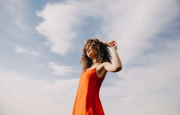 A woman in an orange dress poses against a cloudy sky.