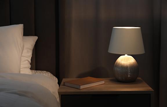 A lamp with a white shade is on a bedside table next to a book.