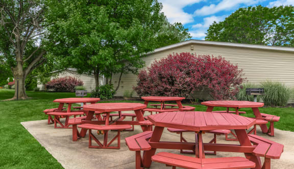 a group of red picnic tables in a grassy area with trees and a building in the