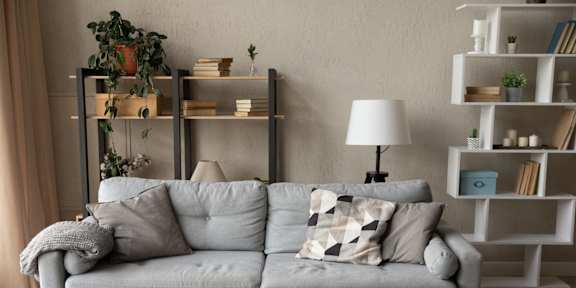A grey couch with pillows and a grey blanket sits in front of a white shelf with books and plants.
