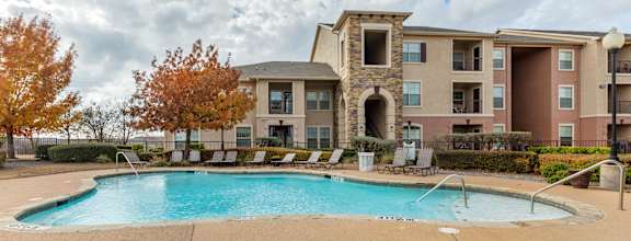 A swimming pool is surrounded by a concrete patio and a building with a stone pillar.