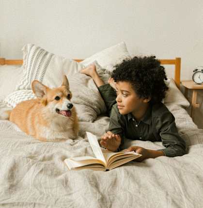 A person is reading a book on a bed with a dog lying next to them.