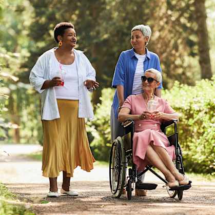 A woman in a wheelchair is being pushed by two women on a path in a park.